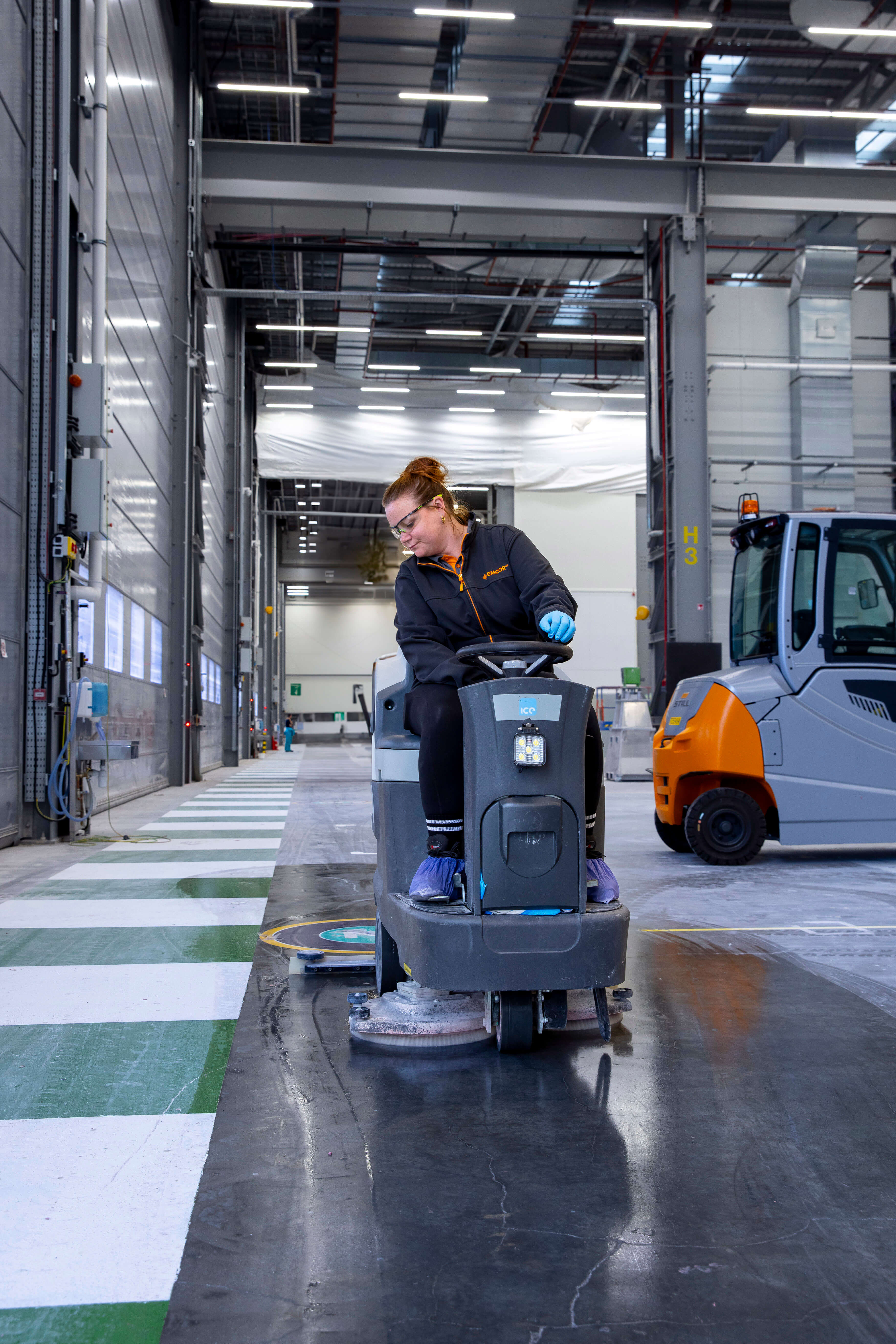 Worker operating a ride-on floor scrubber in a large warehouse with high ceilings and an industrial vehicle in the background.