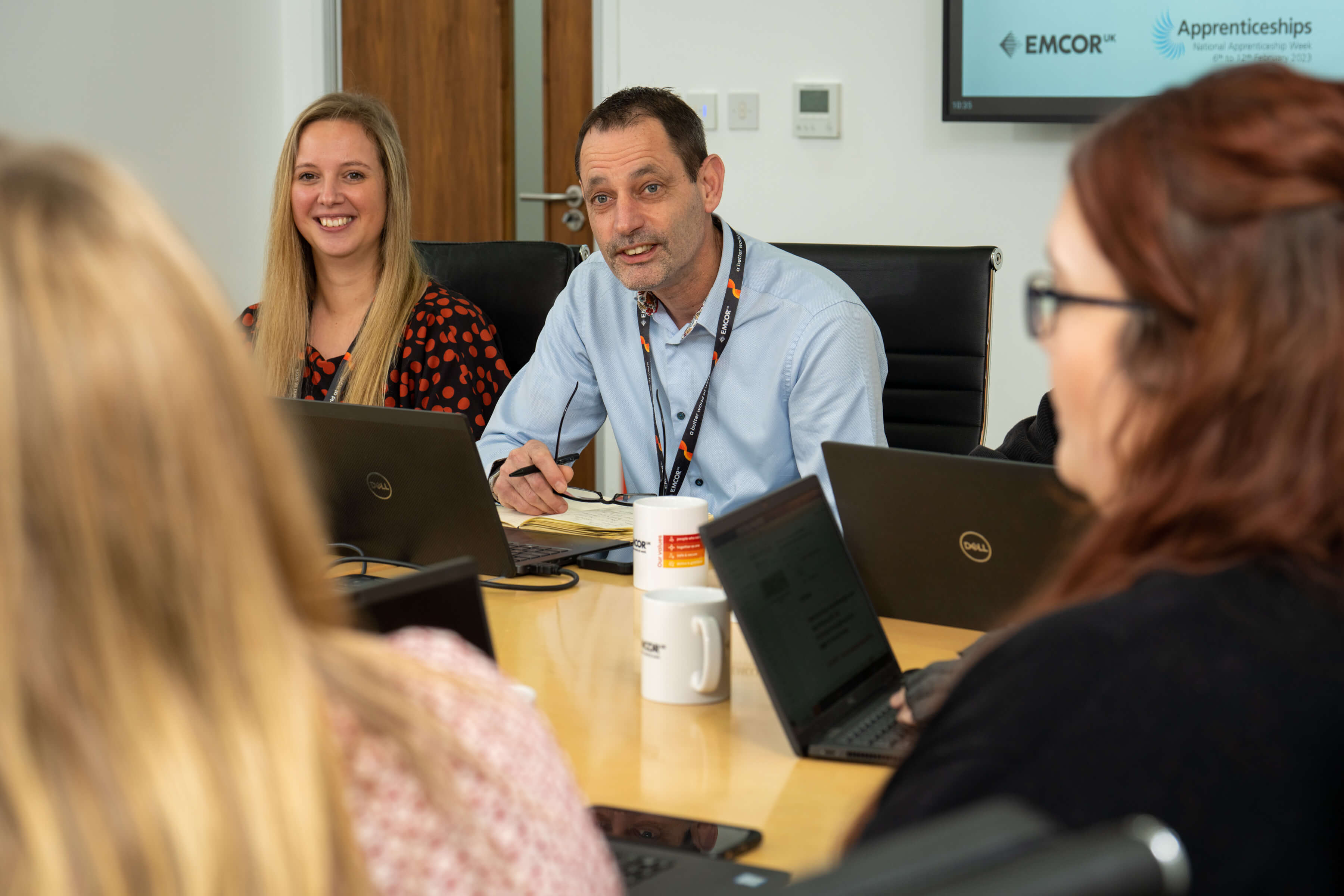 Four colleagues sitting in a meeting room with laptops on the desk. Corner of a PowerPoint showing in the background. 