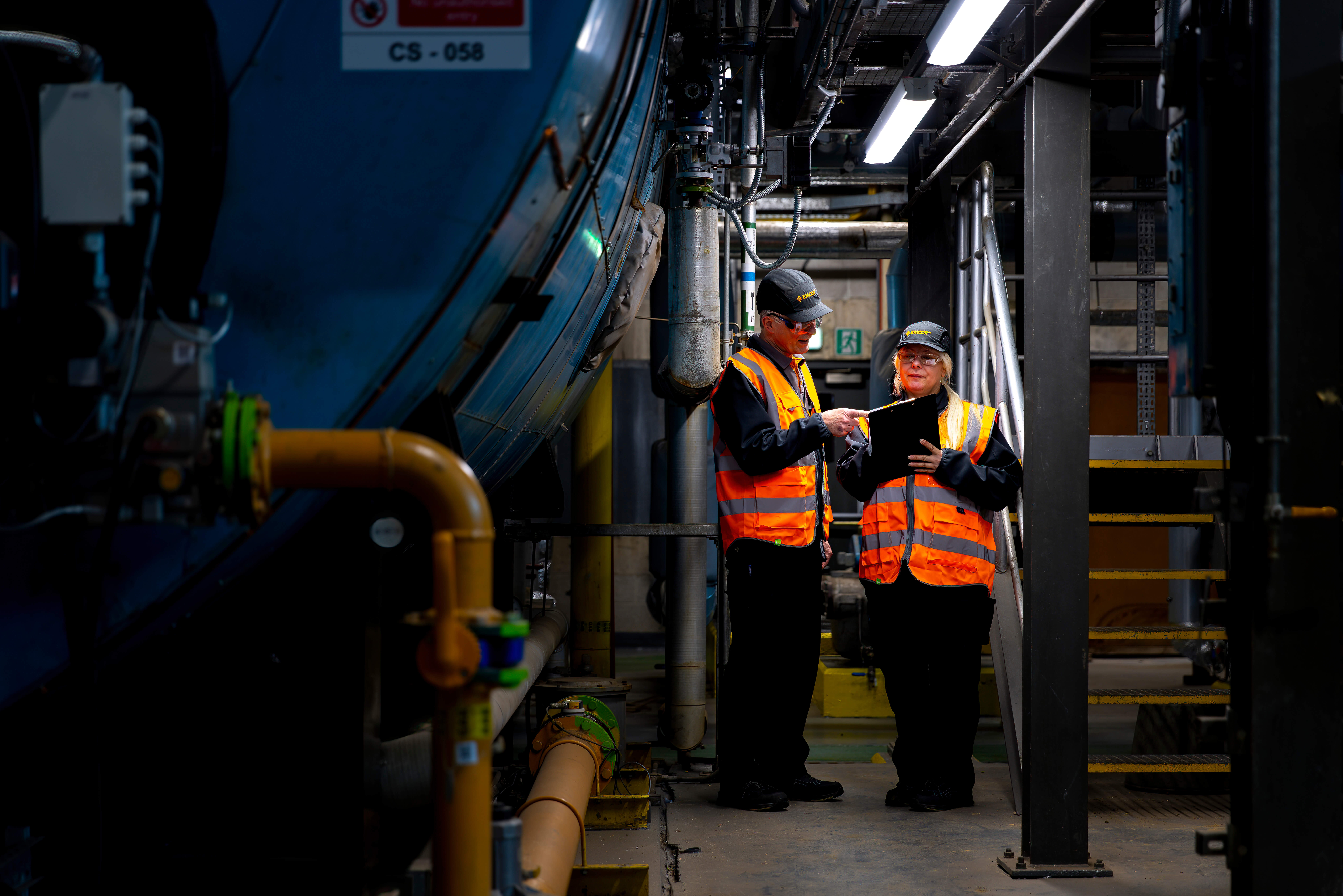 Male and female engineers standing in a boiler room. Female engineer holding clip board while male engineer is pointing at it.