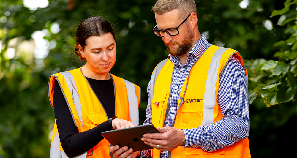 Two EMCOR UK employees wearing safety gear looking at a tablet