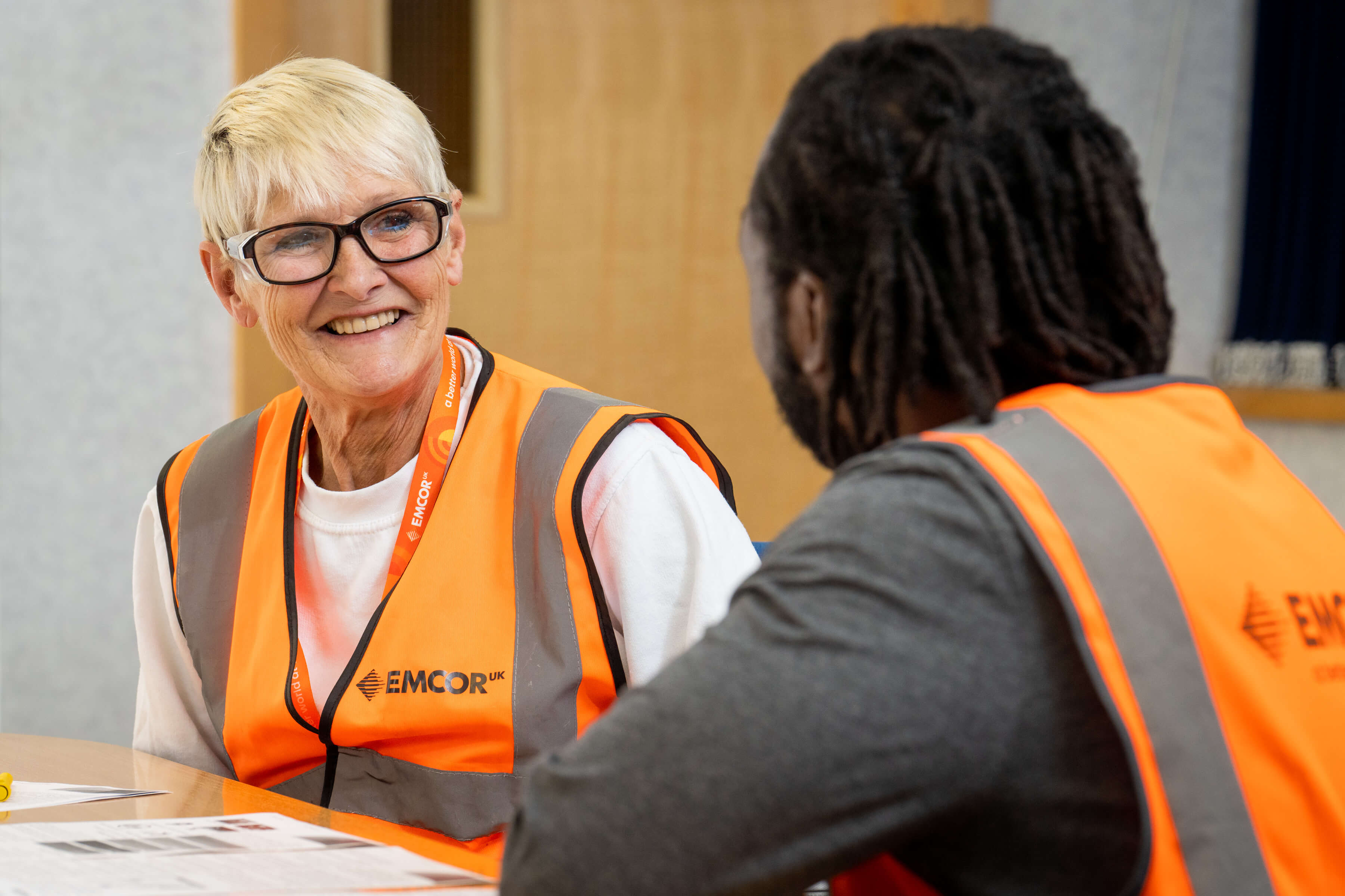 Male and female colleagues wearing EMCOR UK high vis talking in a meeting room. 