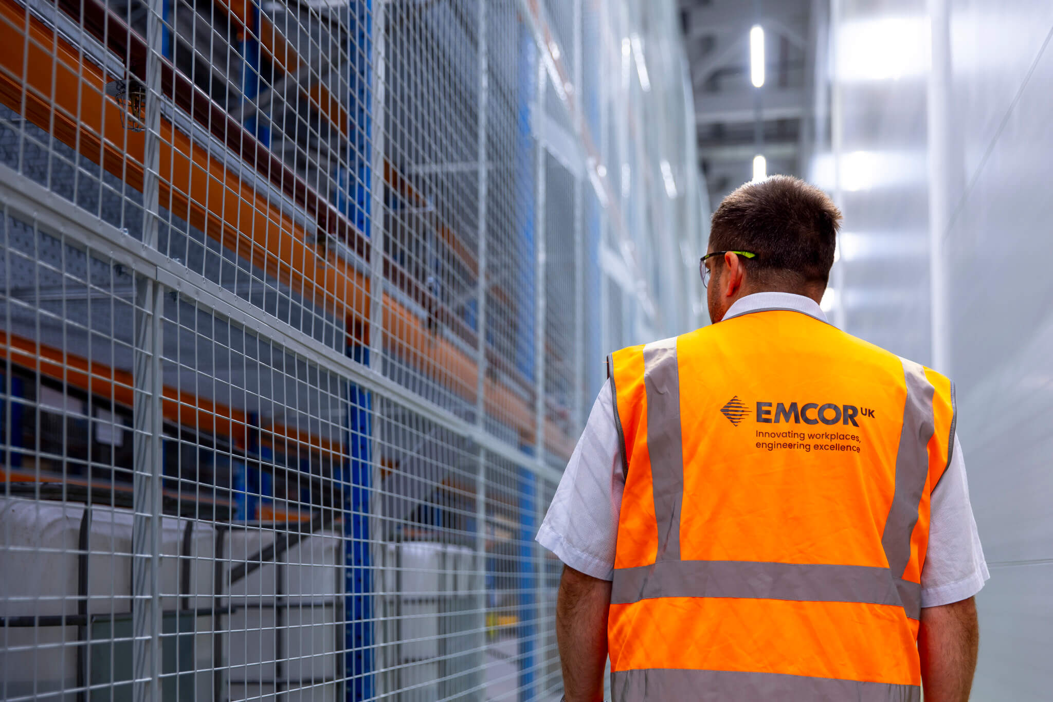 Person walking down a warehouse corridor wearing short sleeve shirt and orange high vis.