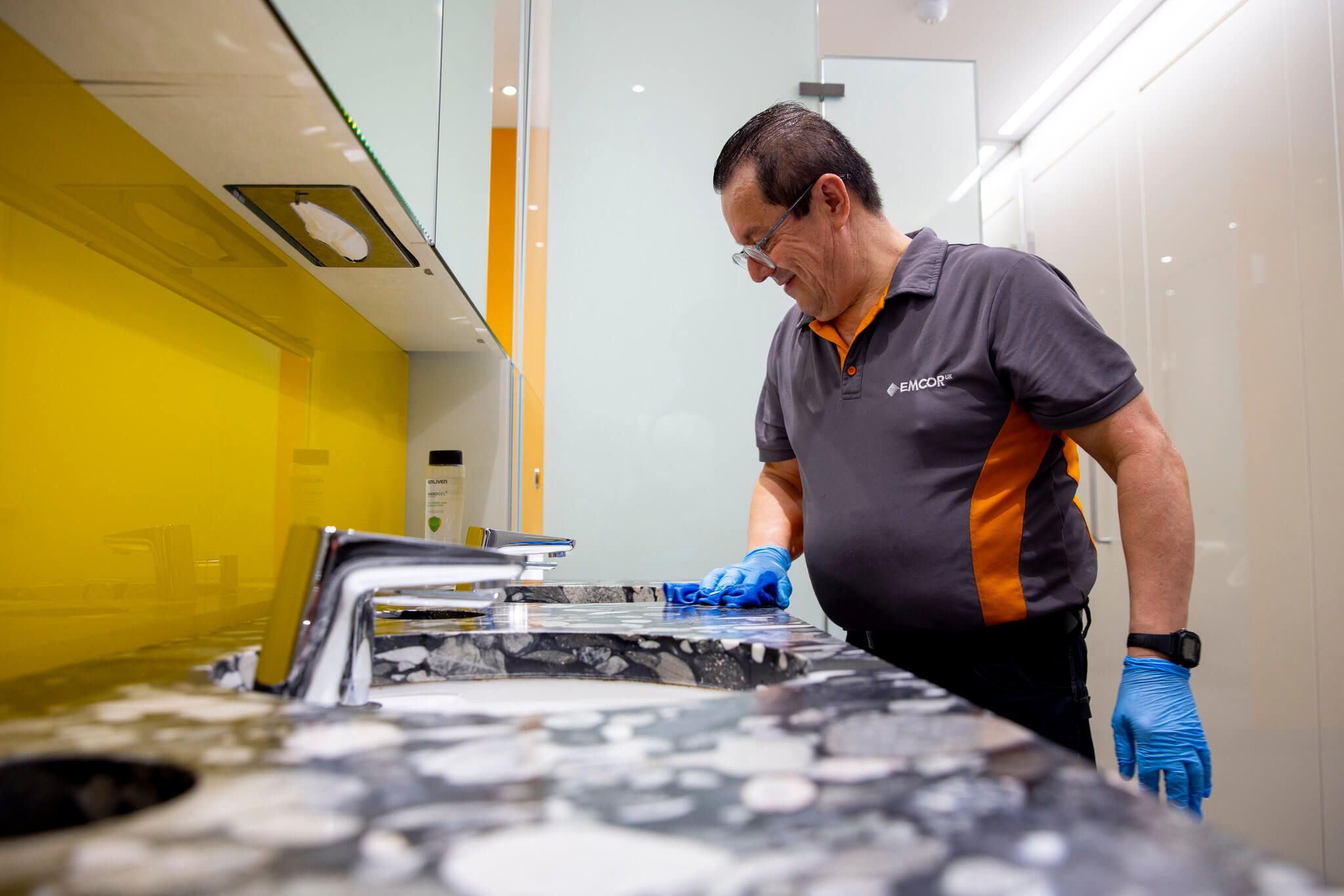 Male cleaner wearing blue gloves in corporate office toilet wiping sinks. Yellow backsplash and cubicles in the background. 