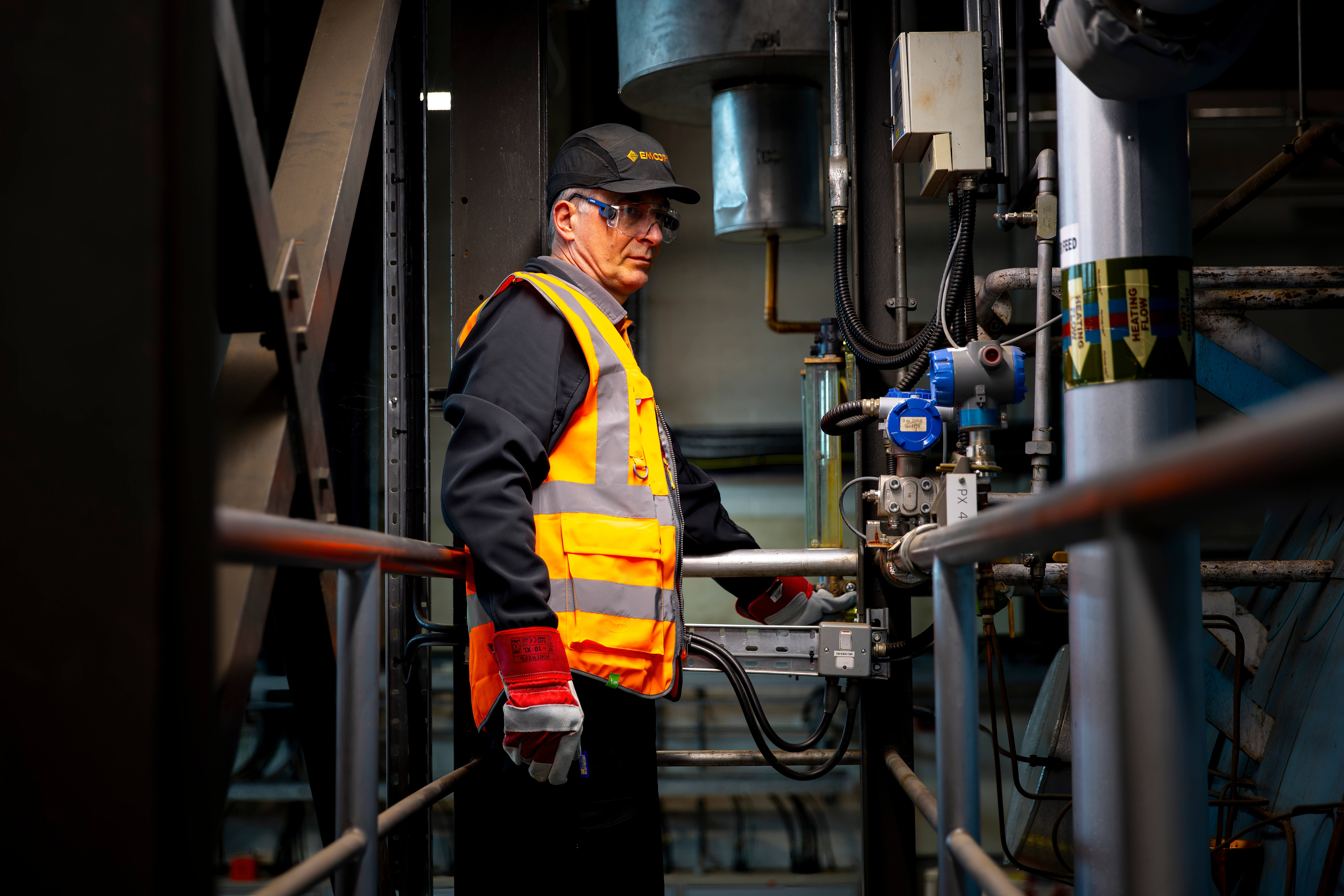 Man in an orange EMCOR UK safety jacket in a boiler room. Handrails and pipes in the surrounding environment .