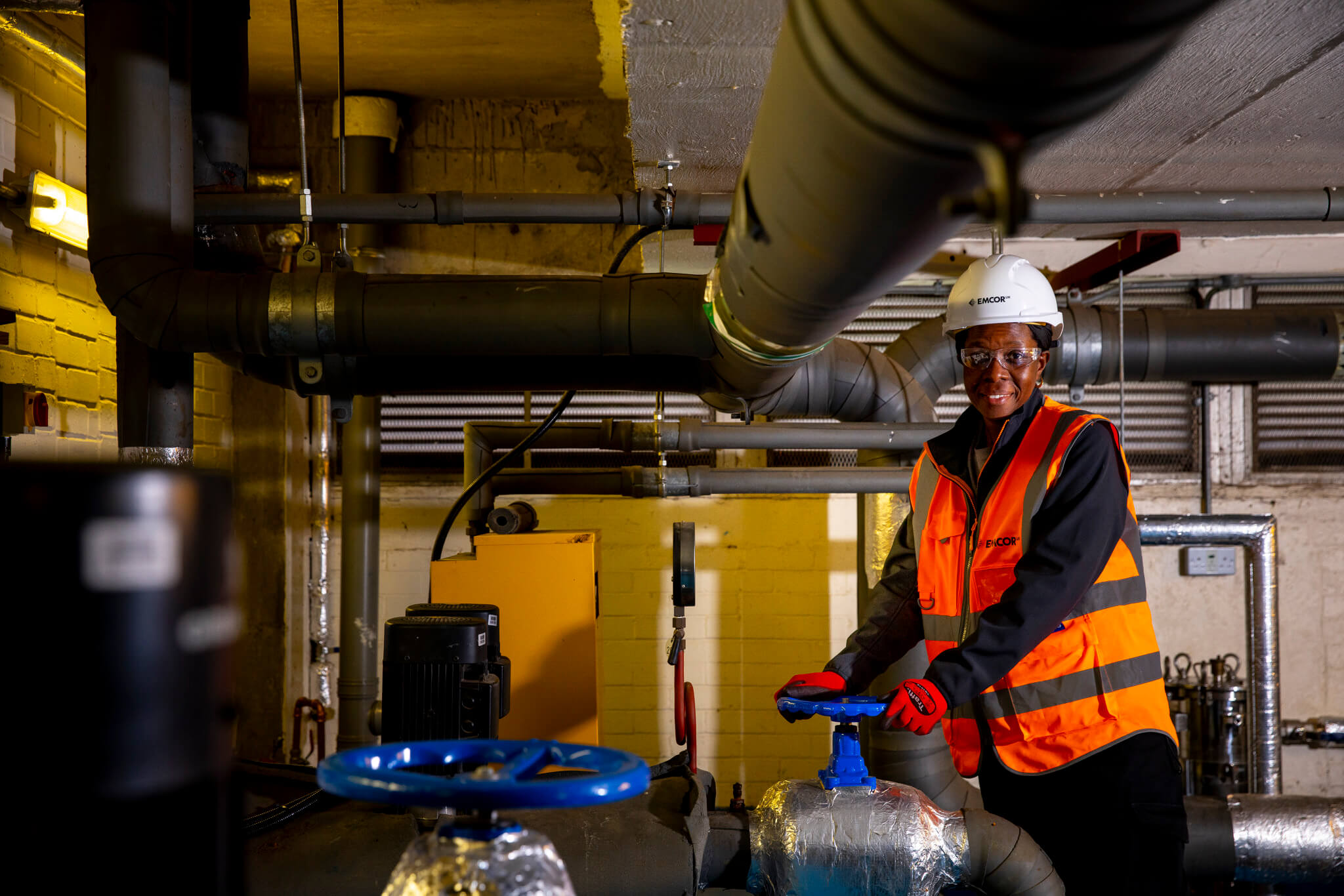 Female engineer in orange high vis, glasses, hard hat and gloves stood in dark boiler room holding water valve.