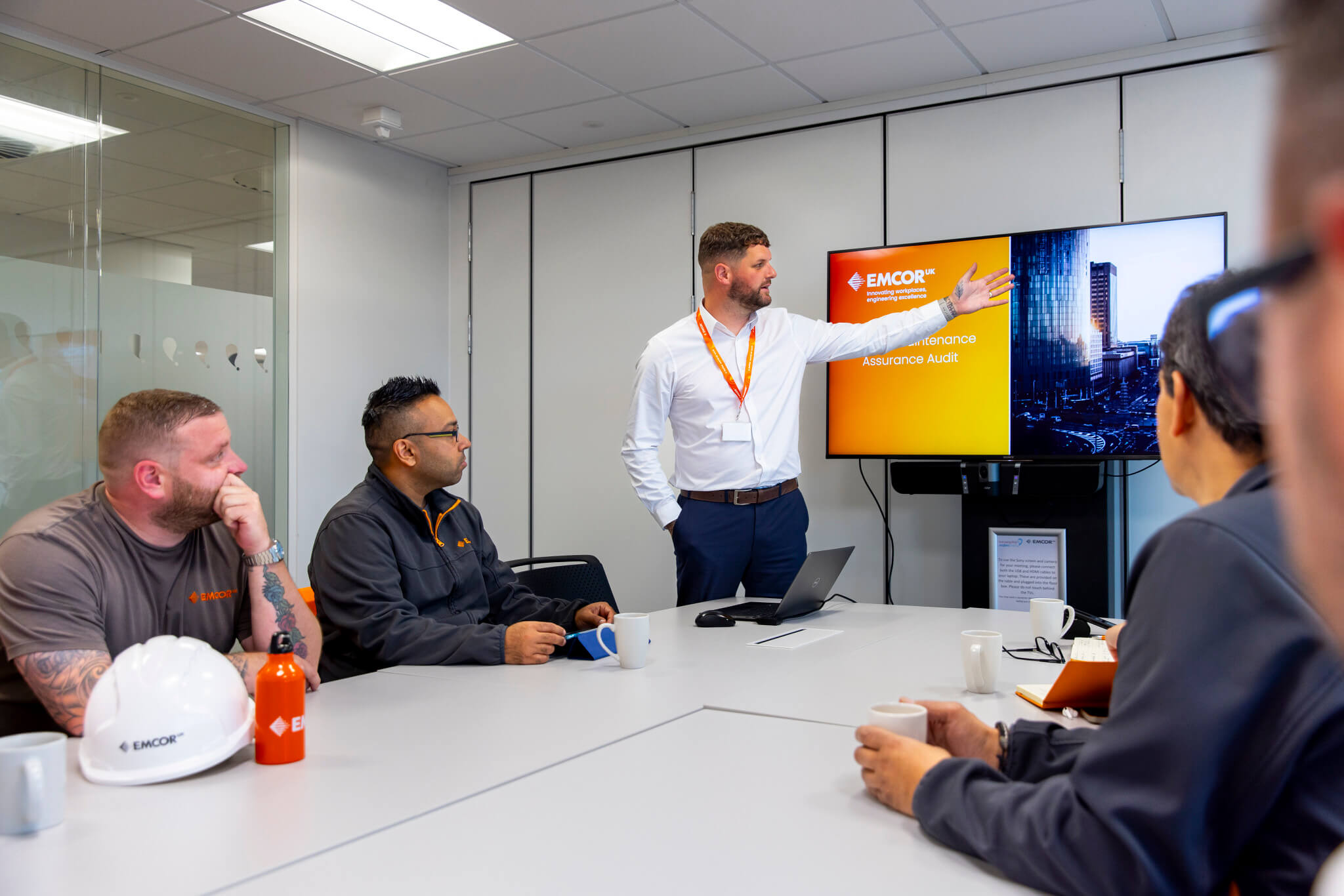 Four people listening to male presenting on TV in a meeting room. EMCOR UK hard hat and water bottle sitting on the desk. 