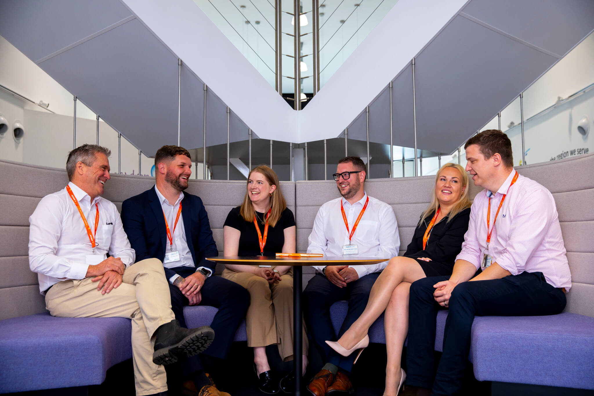 Six colleagues sat in seating area in an office. All wearing orange lanyards wearing formal wear. Staircase in the background. 