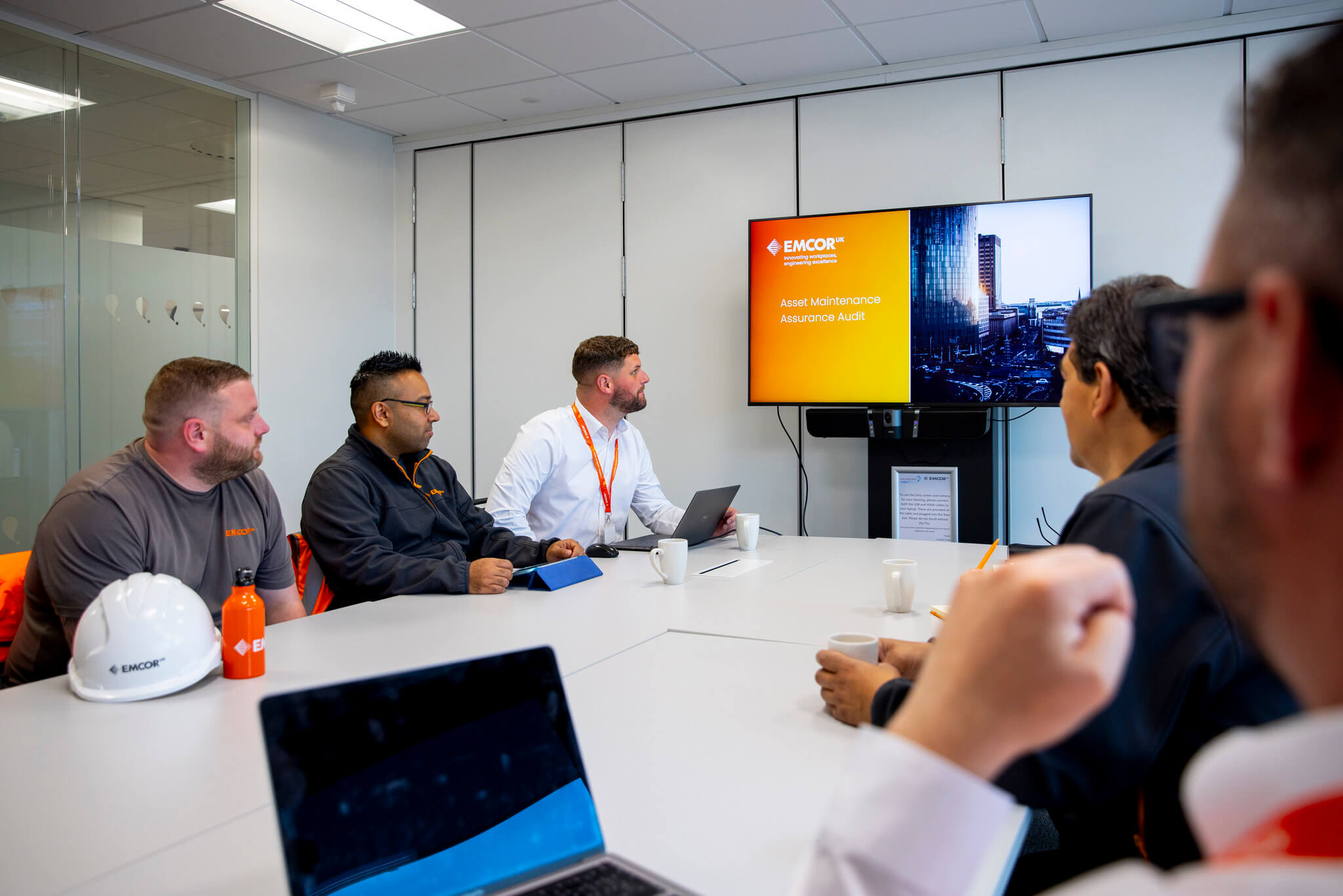 Five colleagues sat in a meeting room with personal items on the desk. Looking at a presentation on large TV.