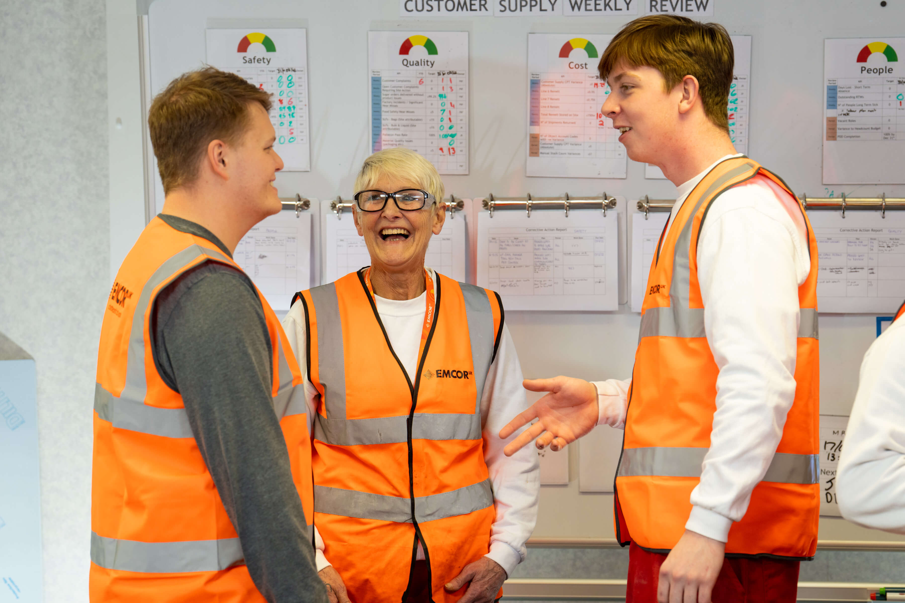 Three colleagues wearing orange high vis stood talking in front of notice board. 