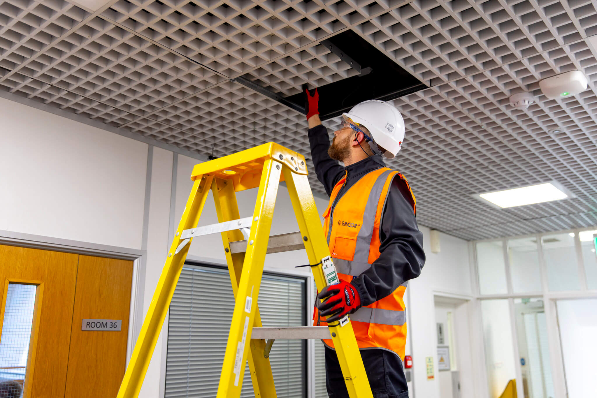 Engineer wearing orange high vis and helmet on a ladder replacing faulty indoor ceiling tiles