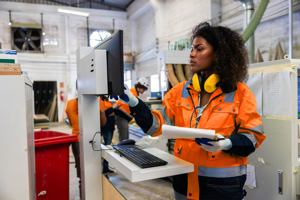 Female technician in high vis jacket working at a manufacturing station, with other workers behind her 