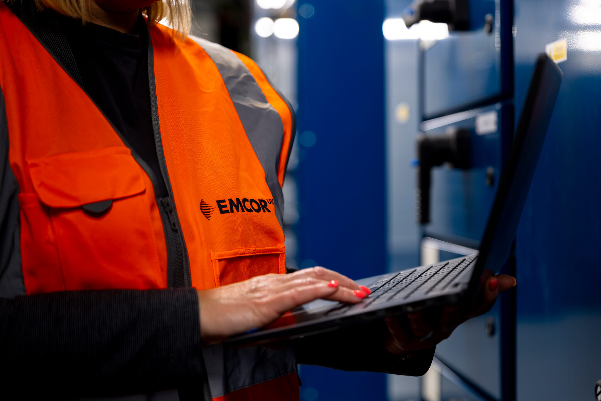 Female technician holding and working on laptop. Blue doors in the background with industrial lighting.