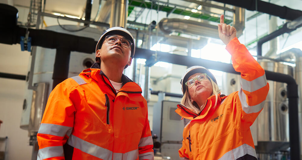 An EMCOR UK female employee and a male apprentice wearing orange EMCOR UK jackets look up to discuss a project in a facility with piping and large equipment.