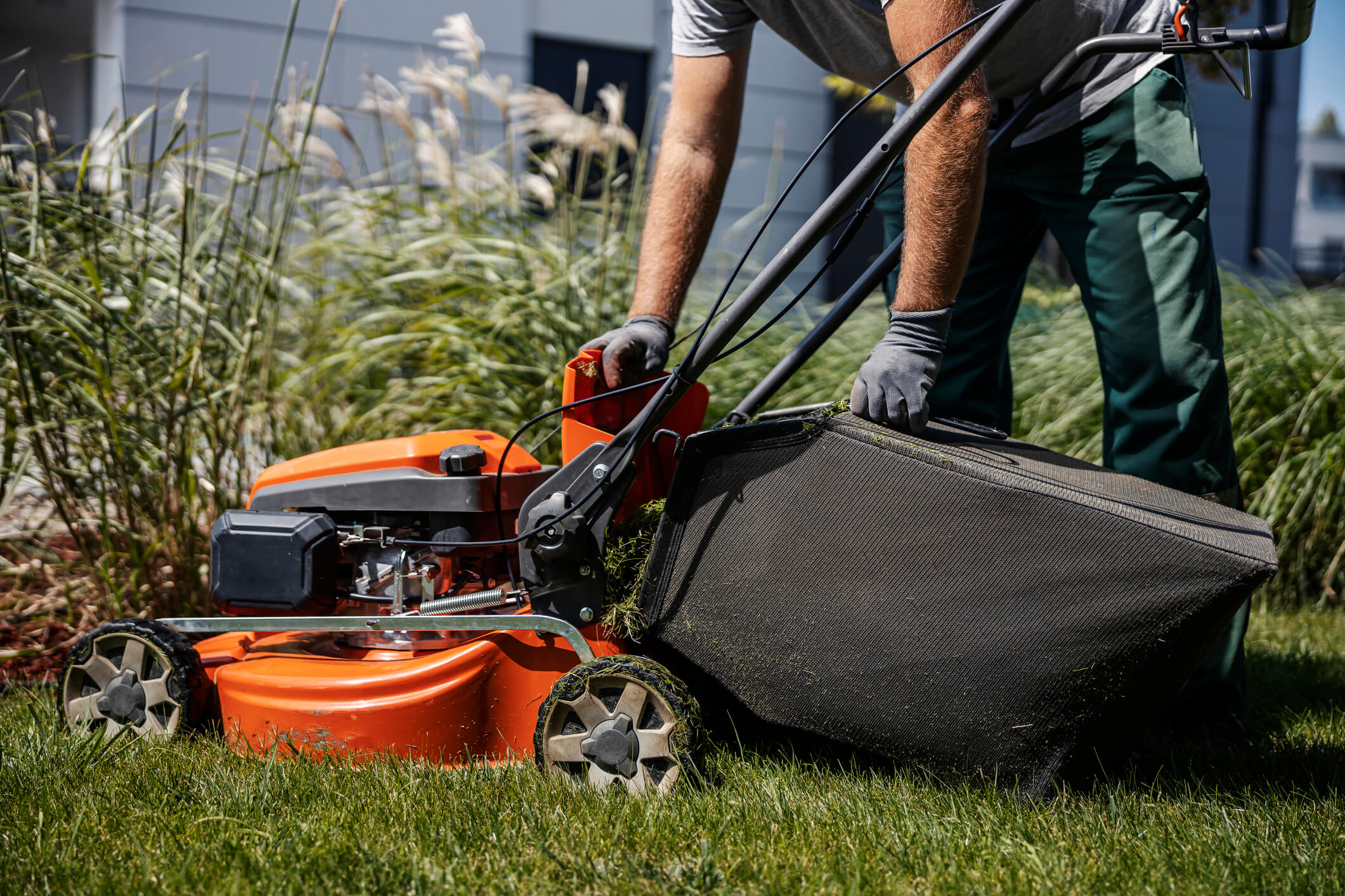 Close up of a gardener wearing grey gloves and t-shirt on grass outside office building. Reaching down to orange lawn mower. 