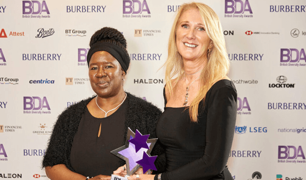 Two colleagues in black dresses holding an award in front of British diversity award background.